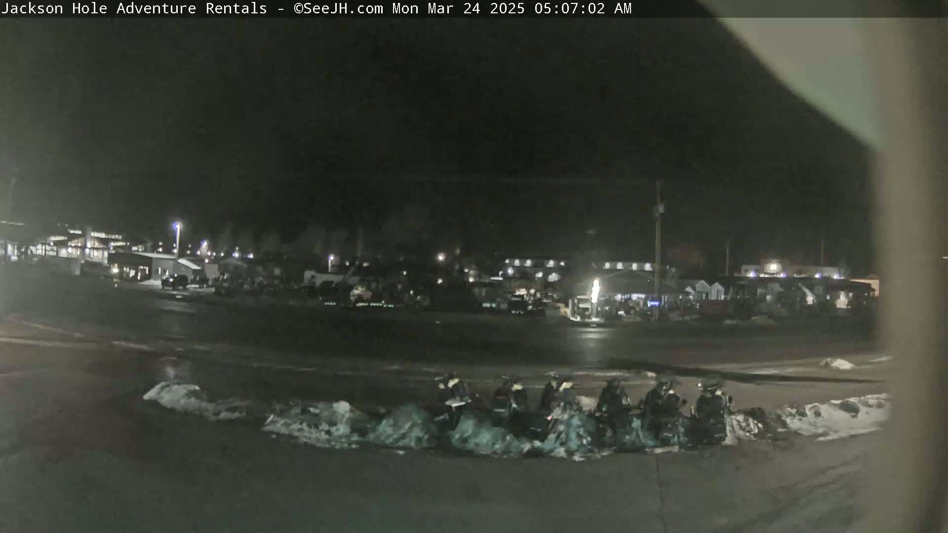 At night, several snowmobiles are parked in a snow-covered lot near a dark building complex.