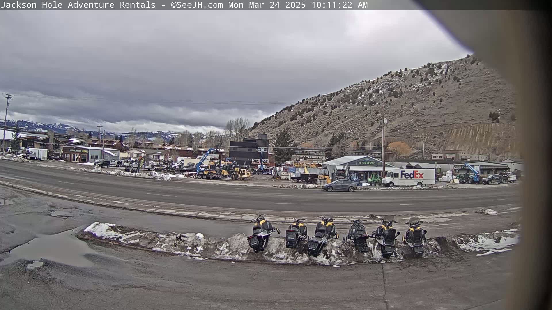 Several snowmobiles are parked near a road in front of a business area with construction equipment and vehicles, under an overcast sky.
