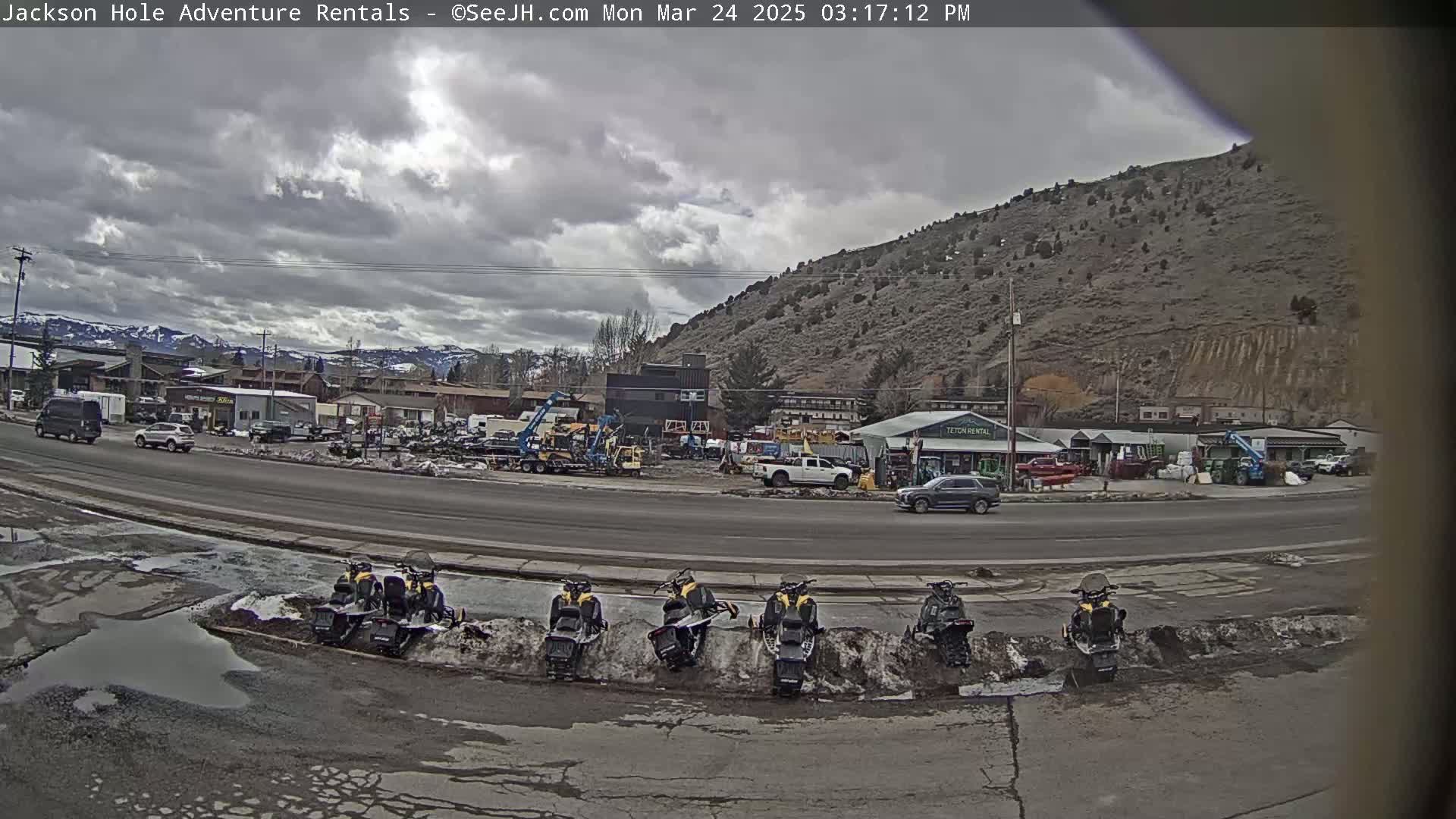 A cloudy day reveals a street scene with several vehicles parked near a row of snowmobiles, in front of a business area nestled against a large, rocky hillside.