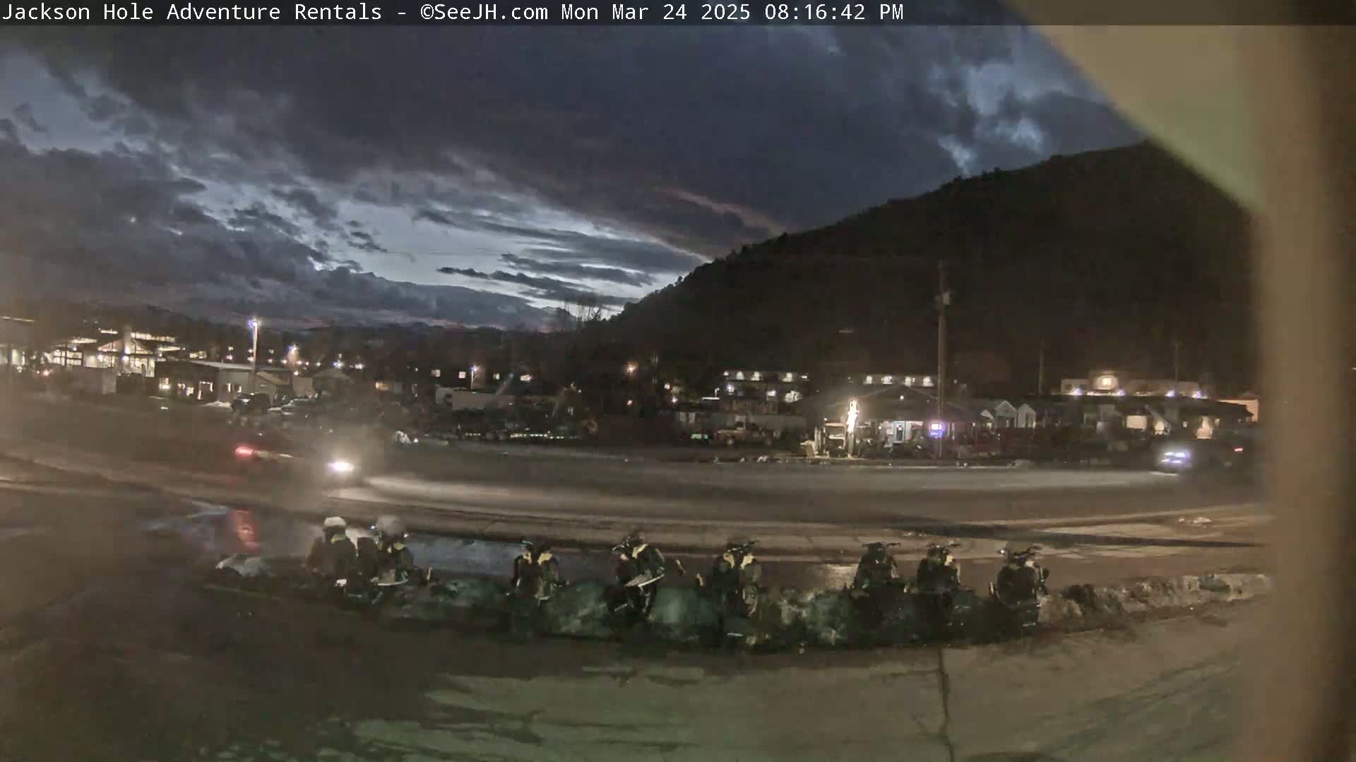 Under a partly cloudy twilight sky, several snowmobiles are parked in a row near a road in front of a town nestled beside a dark mountain.