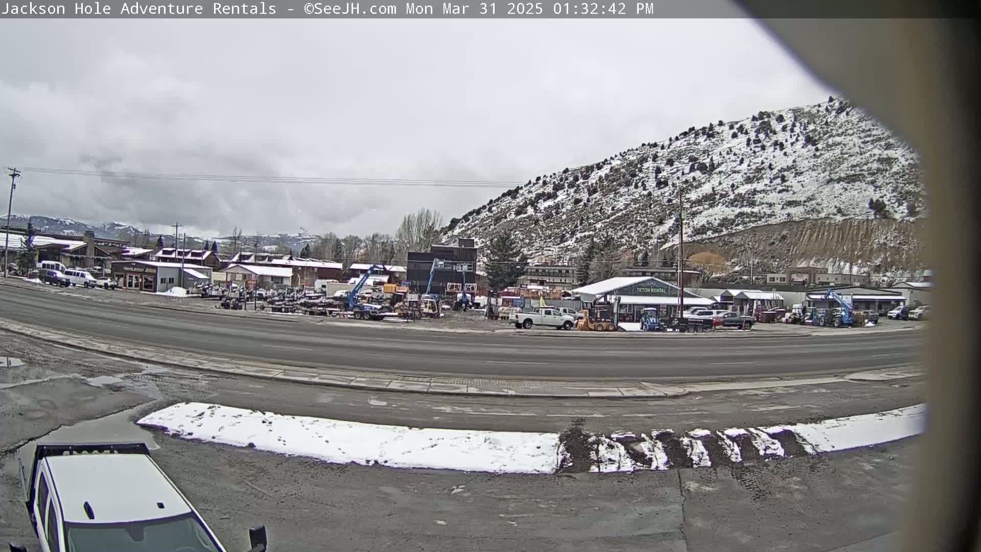 A street scene on an overcast day shows a row of buildings with a snow-dusted hill in the background, along with several vehicles and equipment parked in a lot.