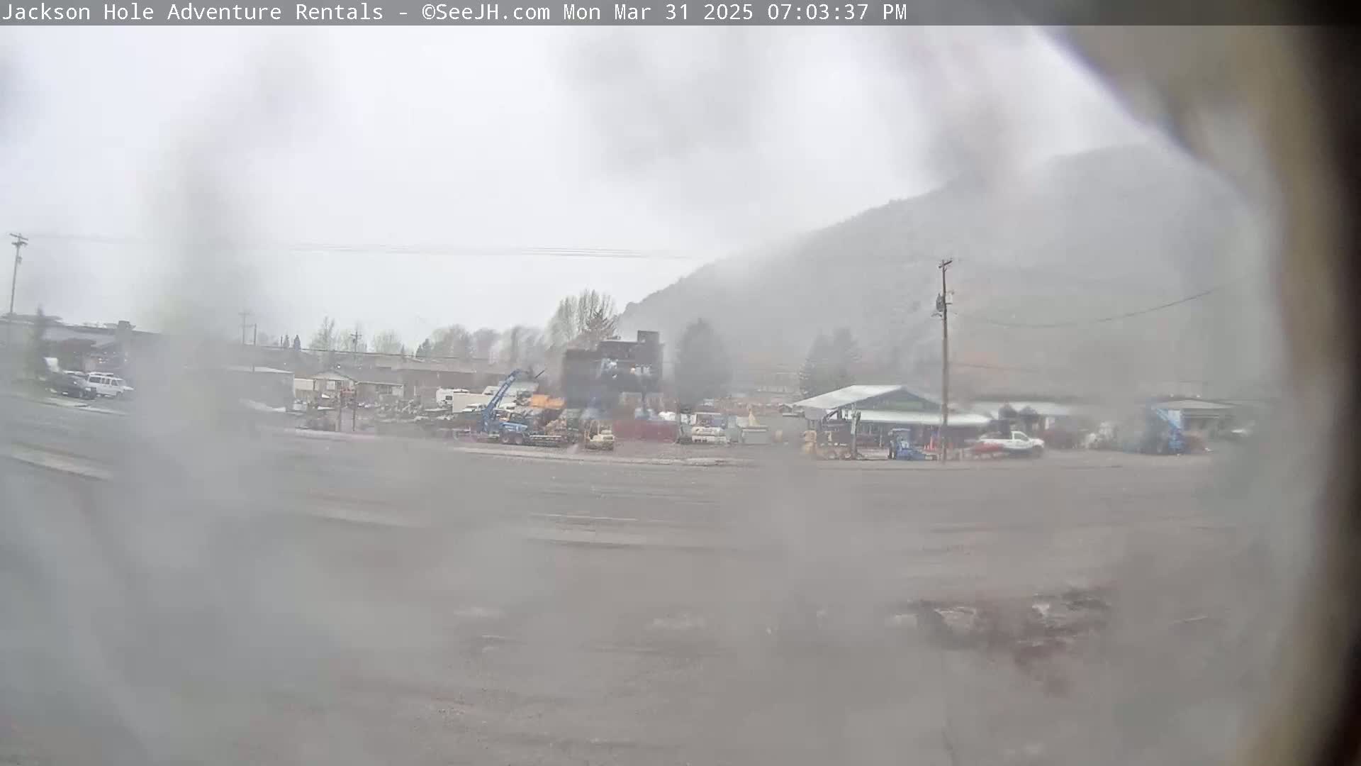 A hazy, rainy scene shows a parking lot with several vehicles and buildings in front of a mountainside.