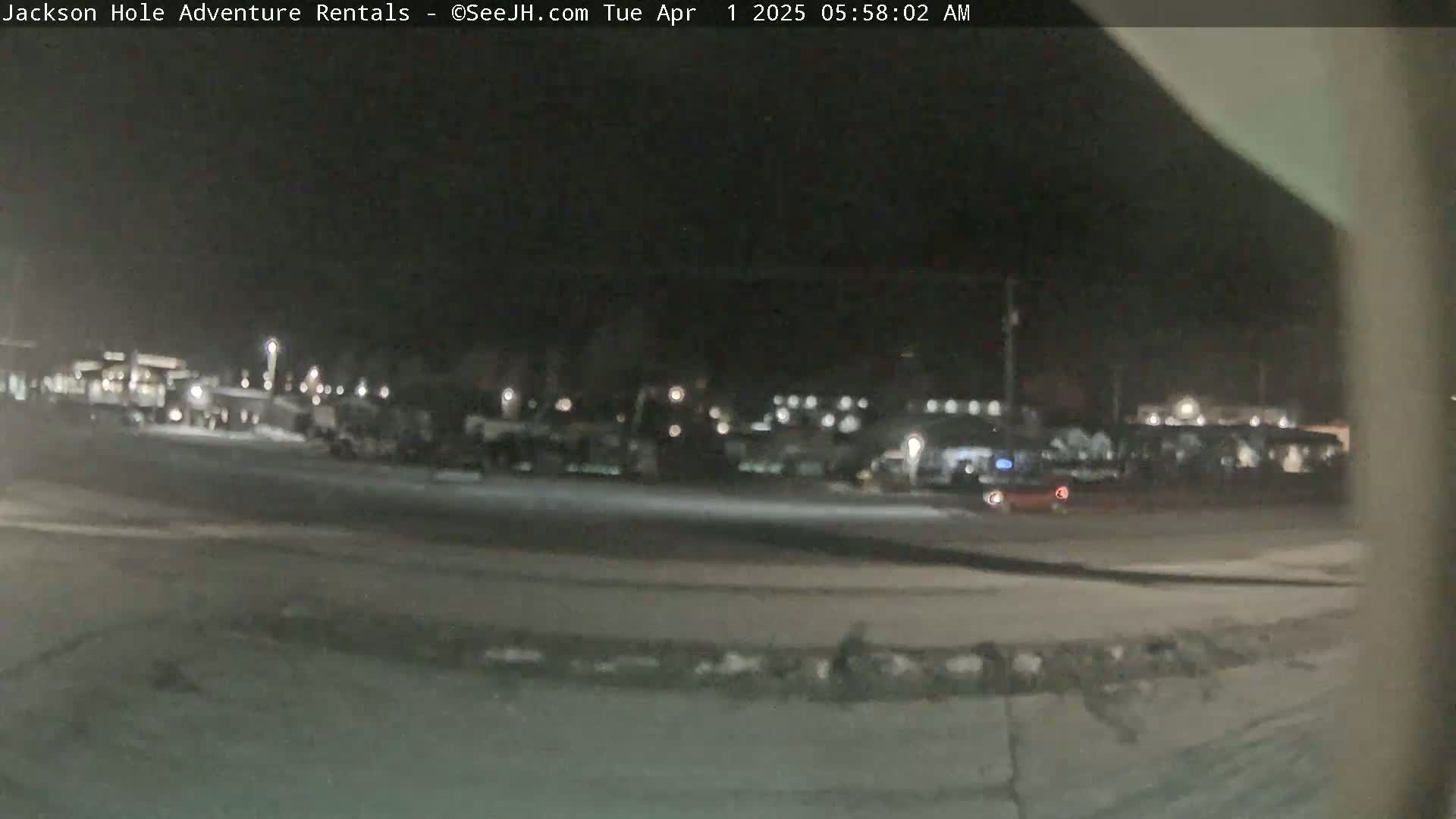 A nighttime, blurry view of a mostly empty parking lot with several vehicles and buildings in the distance.