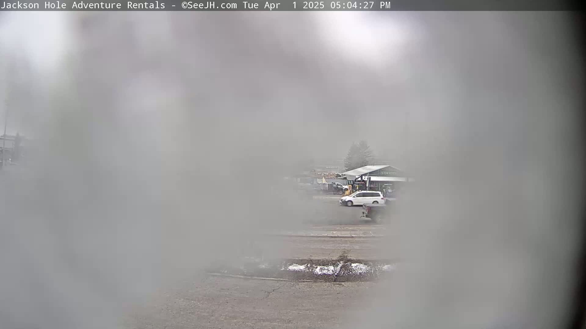 A hazy, possibly snowy or foggy, outdoor scene shows a parking lot with a few vehicles and a building in the distance.