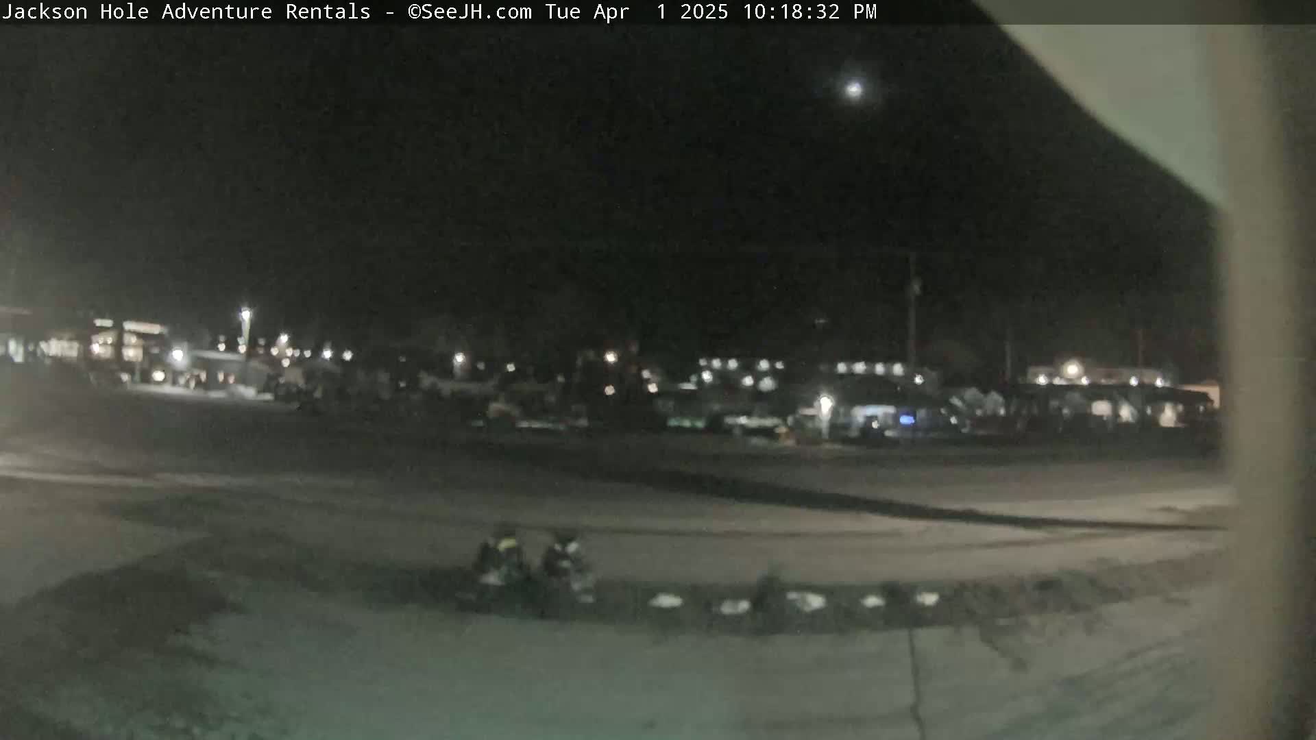 A nighttime, dimly lit outdoor scene shows a parking lot with numerous vehicles and a few people near the bottom of the frame, under a mostly clear sky.