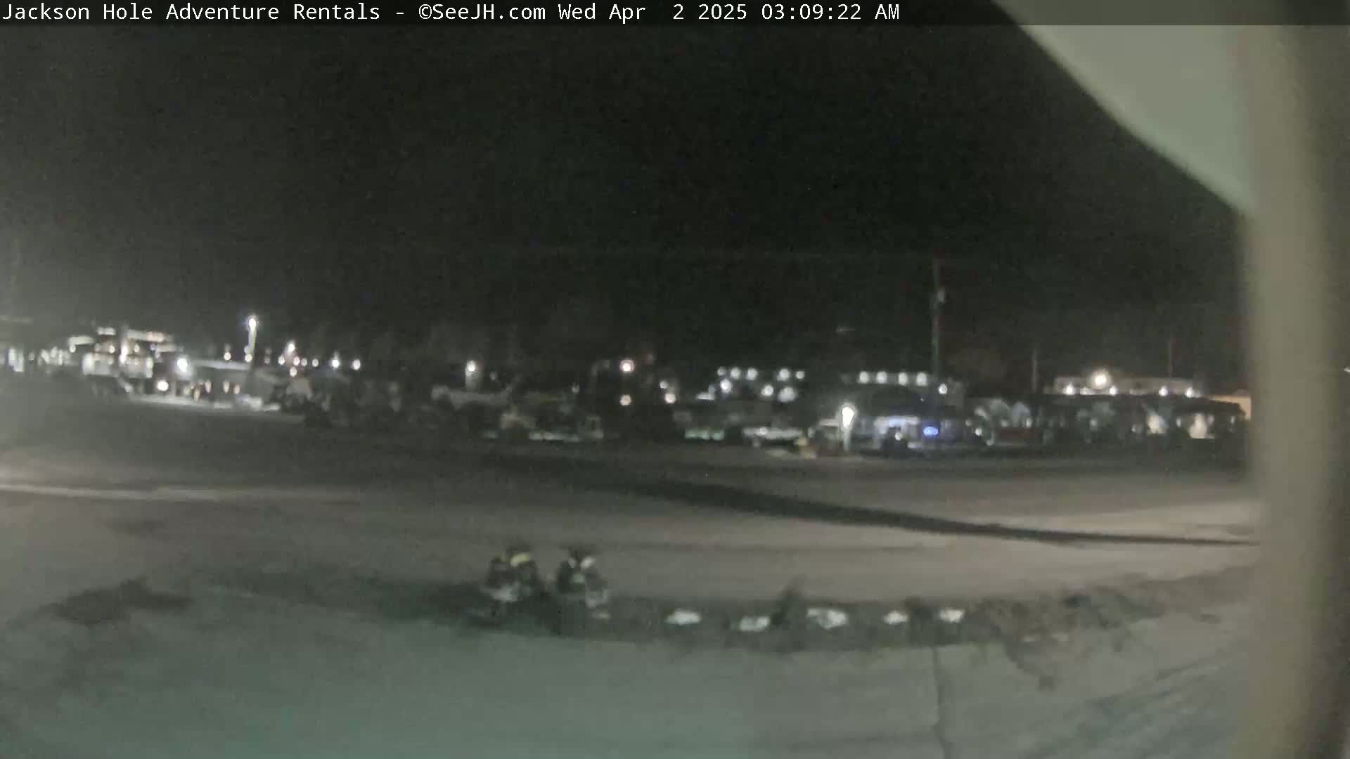 A nighttime, dimly lit parking lot with numerous vehicles and a few figures in the foreground.