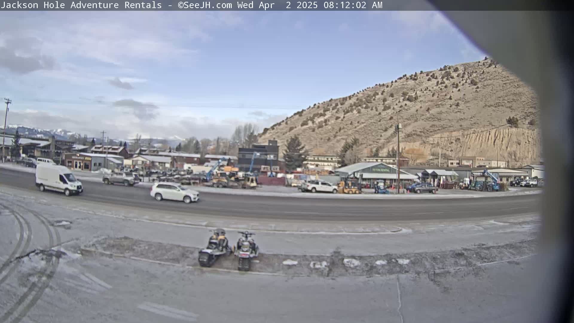 A mostly clear day reveals a street scene with several vehicles, including a white van and SUV,  near a row of buildings and a brown hill in the background, with some patchy snow or ice visible on the ground.