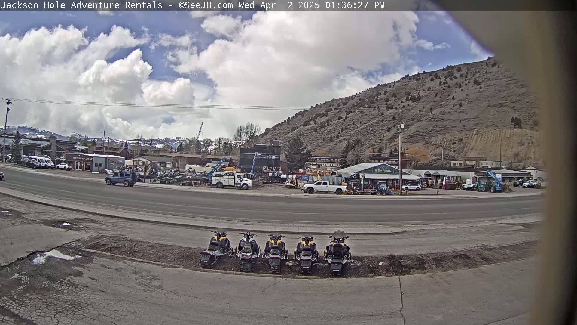 A partly cloudy day reveals a street scene with several vehicles, including a line of snowmobiles parked, in front of various businesses and buildings at the base of a large hill.