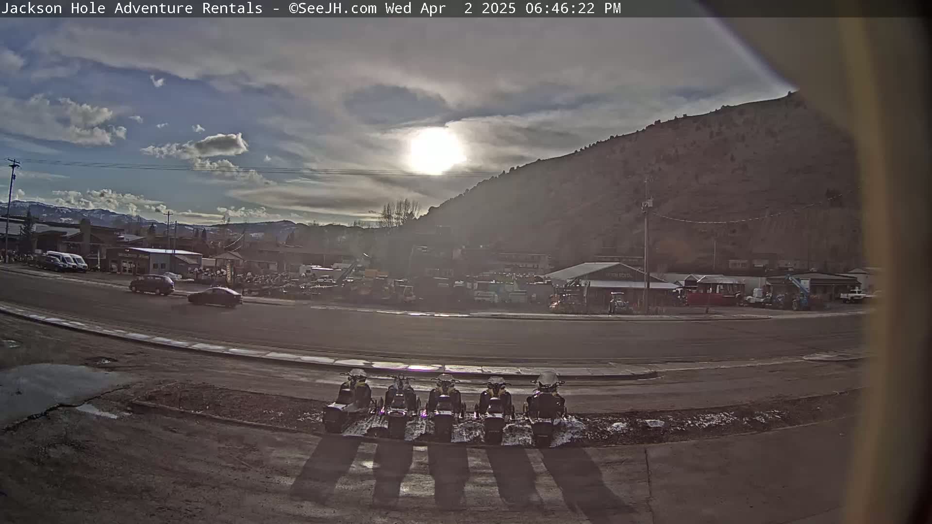 A partly cloudy day reveals a parking lot with several snowmobiles parked in a row, in front of a street with cars and buildings backed by a large hill.
