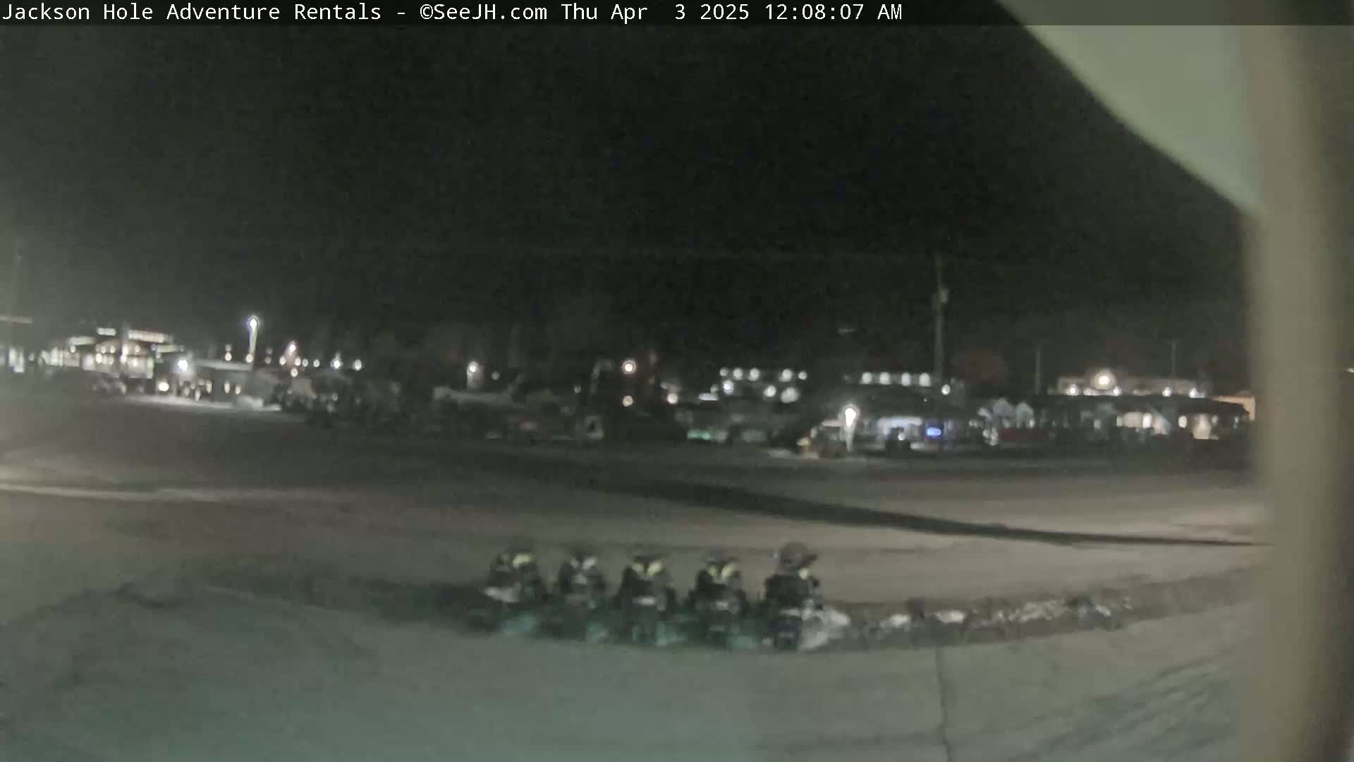 At night, a parking lot with several vehicles is visible, and a row of small, dark vehicles are parked in the foreground.