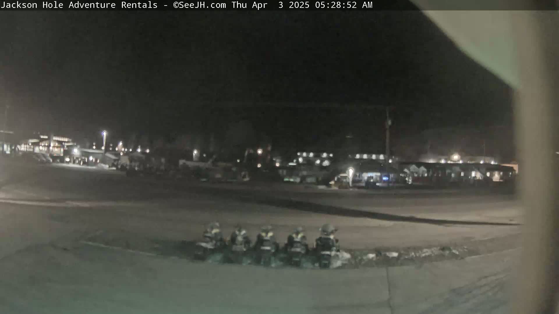 At night, several all-terrain vehicles are parked in a lot near a row of buildings.