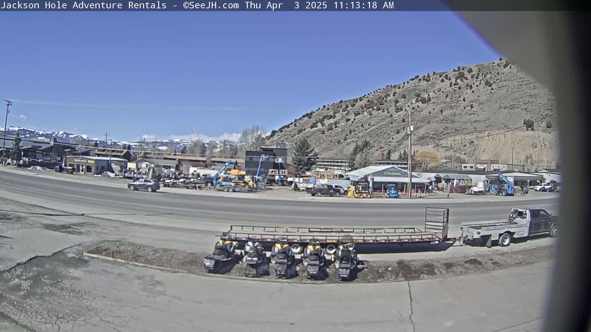 A sunny day reveals a parking lot with several snowmobiles on a trailer, numerous vehicles, and construction equipment near a row of buildings against a backdrop of a hill and distant mountains.