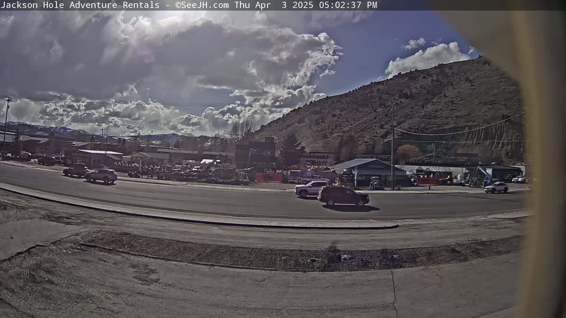 A partly sunny day with fluffy clouds overhead shows a street scene with several vehicles and buildings, including what looks like an equipment rental business, situated at the base of a large hill.