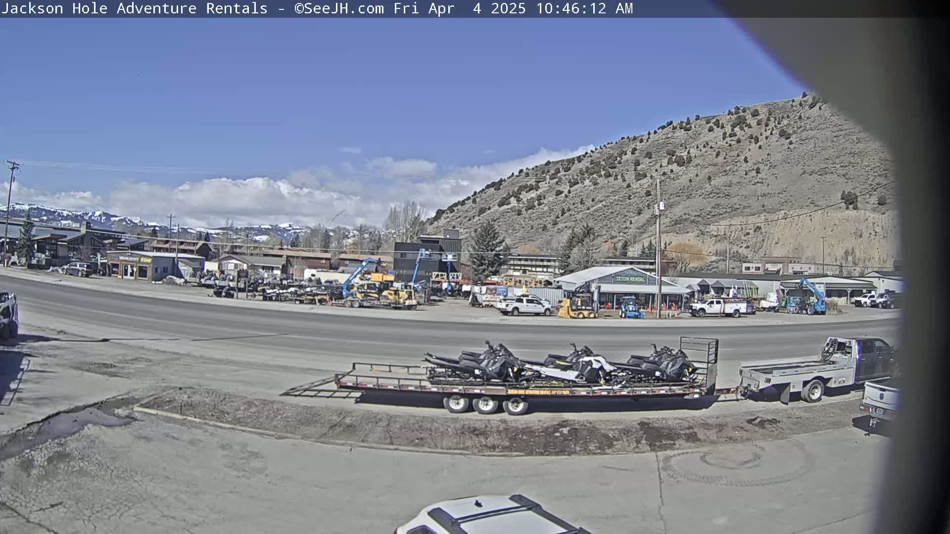 A sunny day reveals a parking lot filled with various vehicles and equipment, including a flatbed trailer loaded with snowmobiles,  in front of a row of buildings nestled against a mountainside.