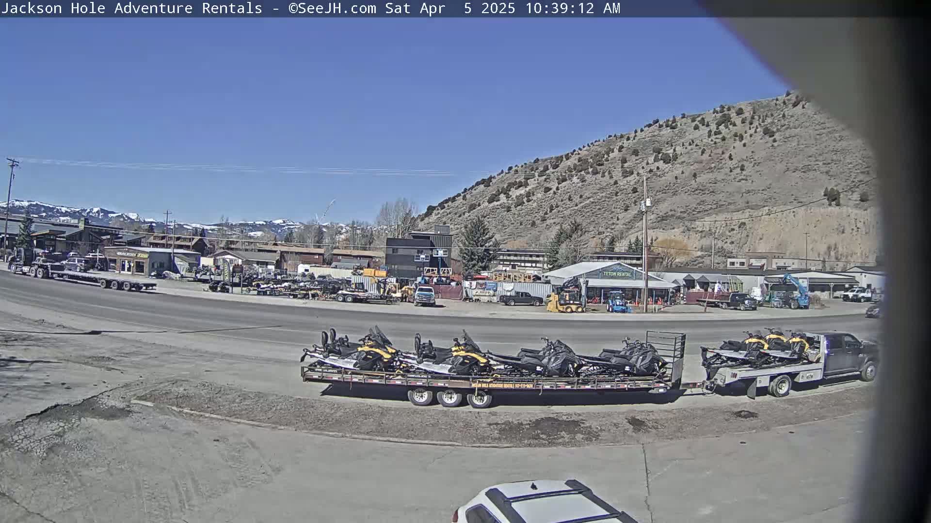 A sunny day reveals a parking lot filled with various vehicles, including a flatbed truck loaded with snowmobiles, and a background of buildings nestled against a hillside.