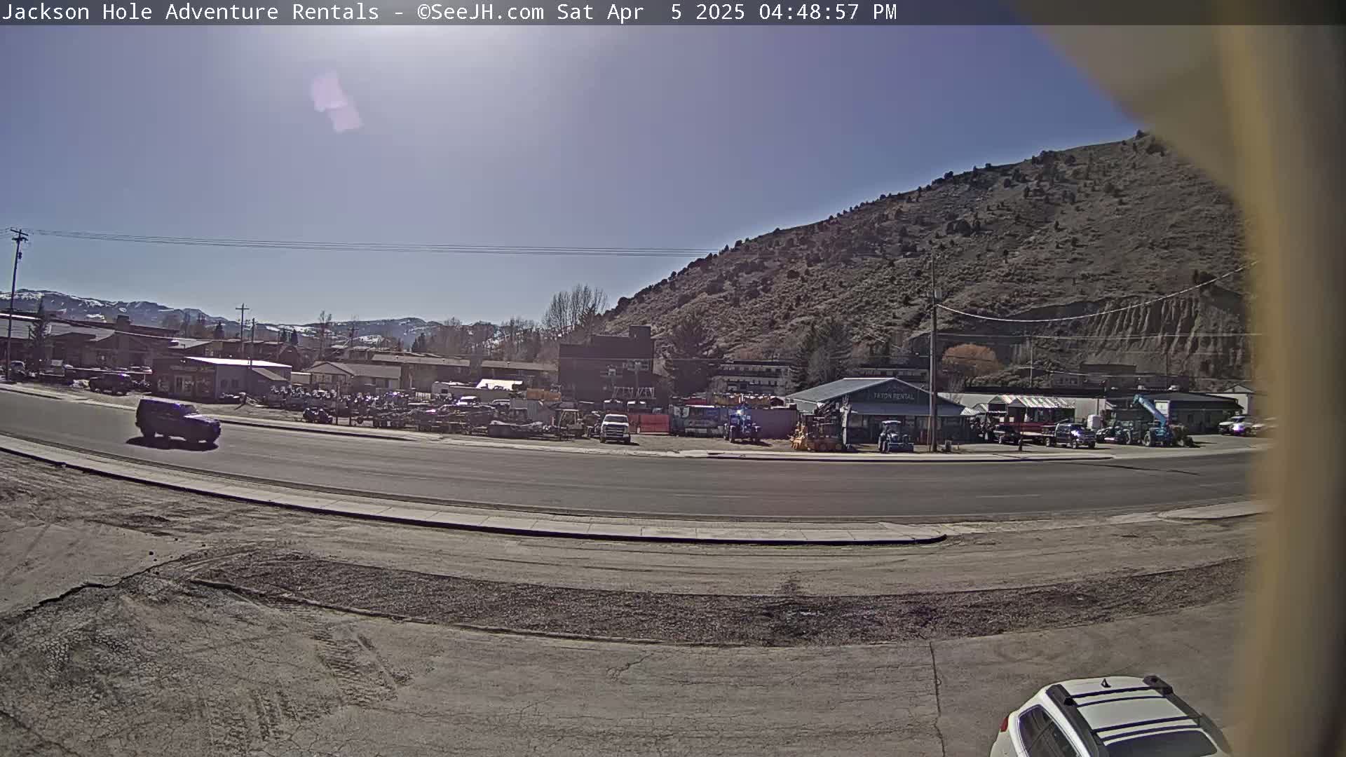 A sunny day reveals a road scene with a few vehicles, a row of businesses, and a large, barren hill in the background.