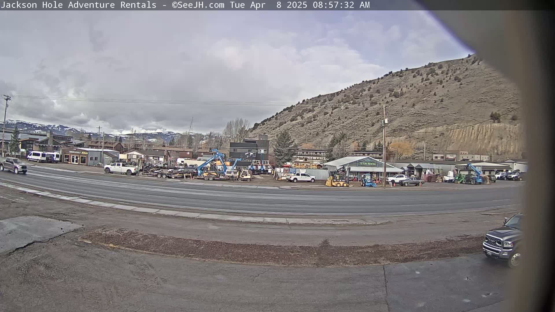 A street scene under an overcast sky shows a row of businesses, some with heavy equipment parked outside, near a hill.
