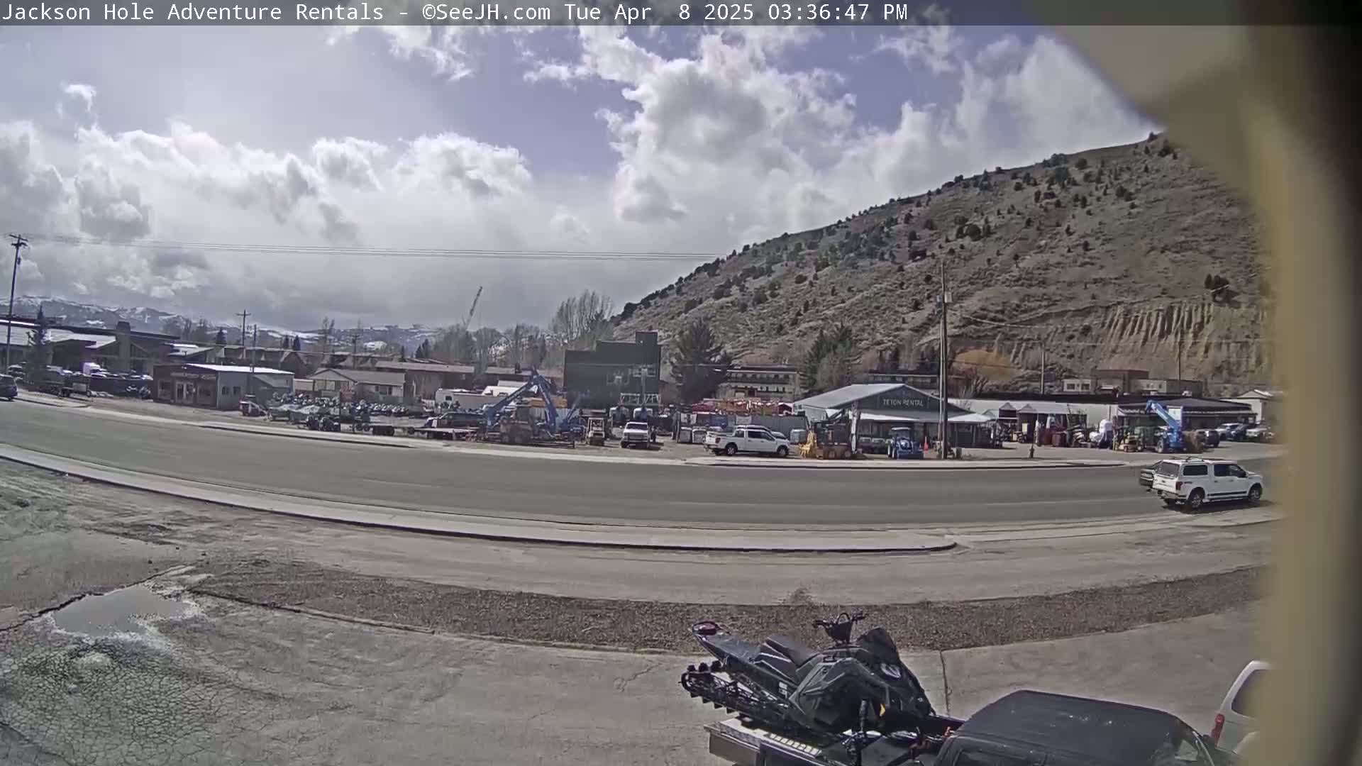A partly cloudy day reveals a street scene with various vehicles, including a snowmobile in the foreground, and several buildings and equipment rentals in the background against a backdrop of a brown hill.