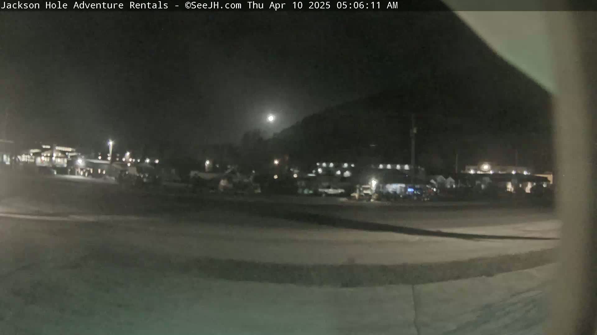 A nighttime, dimly lit outdoor scene shows a parking lot with vehicles and buildings in the background under a bright moon.