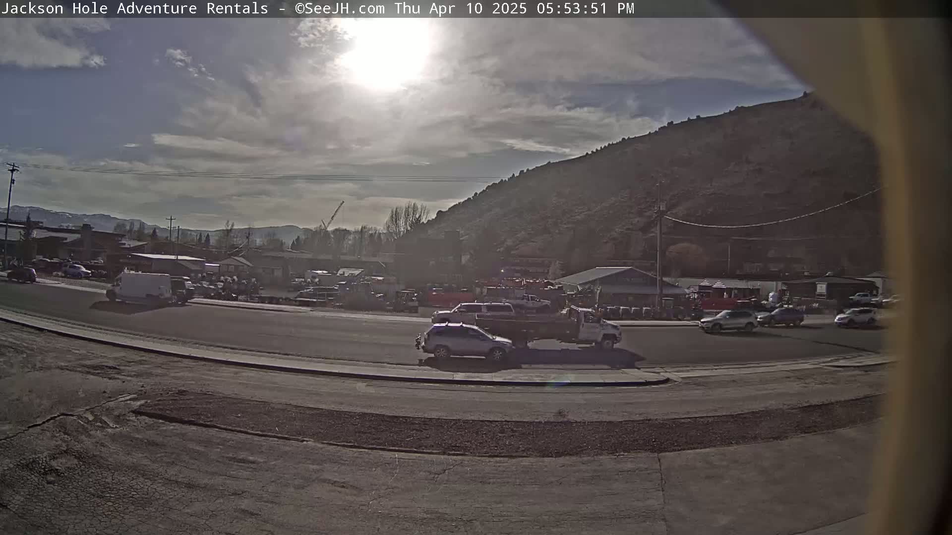 A sunny day with partly cloudy skies shows a street scene with several vehicles, including a tow truck, parked and driving near a hillside in a town with buildings.