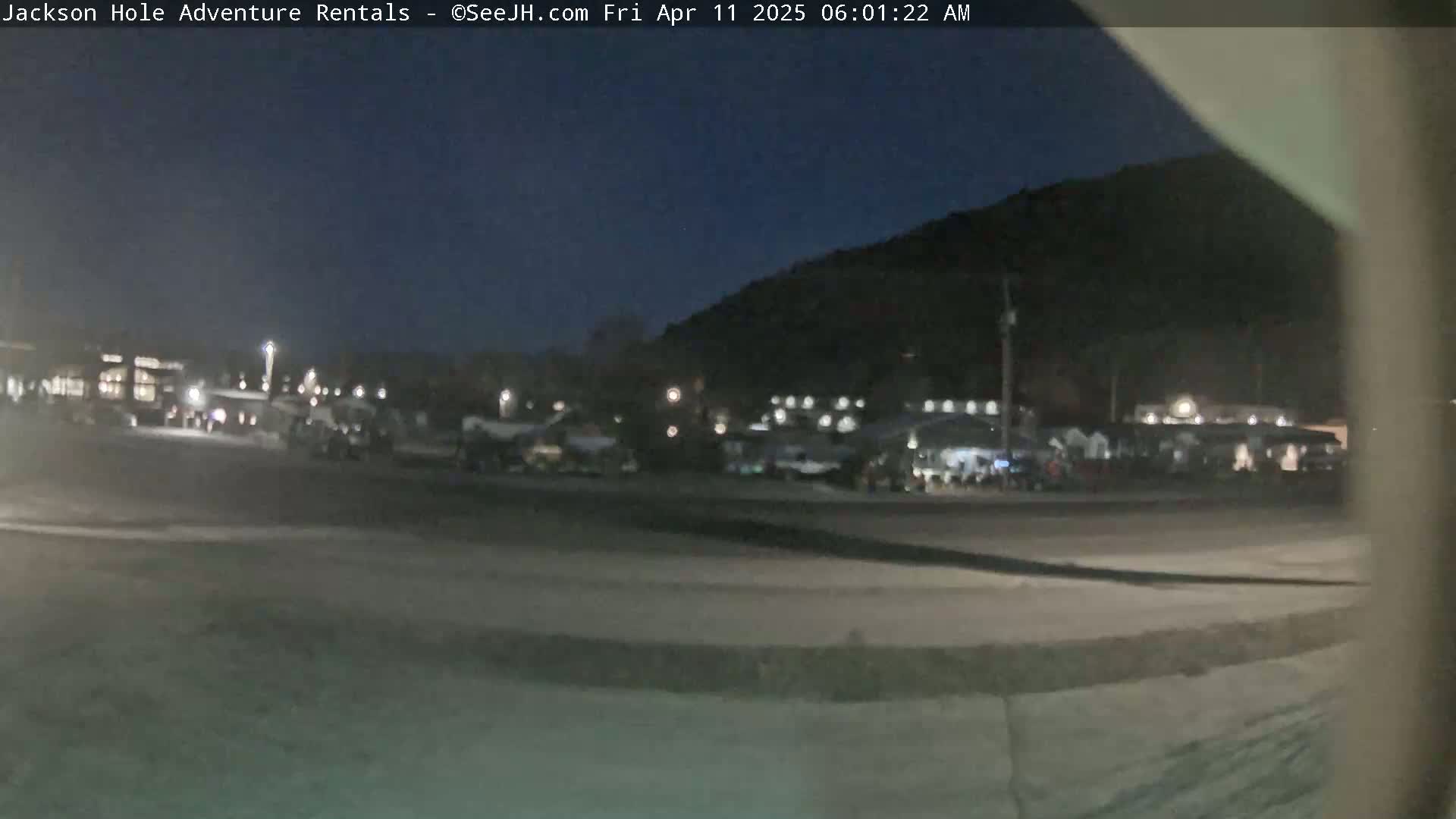 A dark, nighttime scene shows a parking lot with several vehicles, buildings, and a hill in the background under a clear, dark sky.