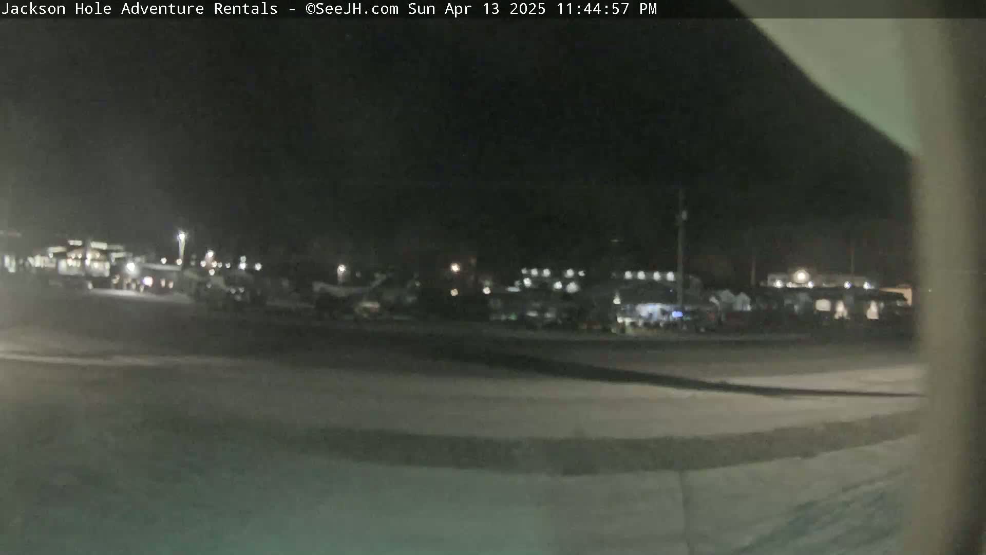 A nighttime, outdoor view shows a large, dimly lit parking lot with numerous vehicles.