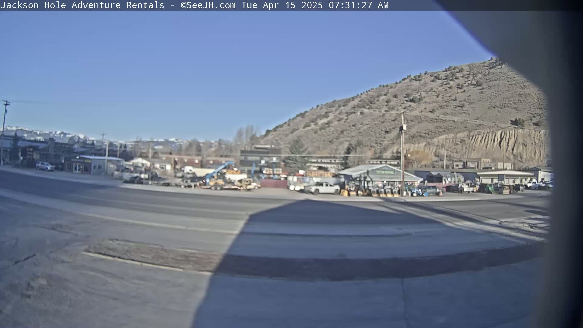 A sunny day reveals a parking lot with various vehicles and equipment near buildings nestled at the base of a large, dry hill.
