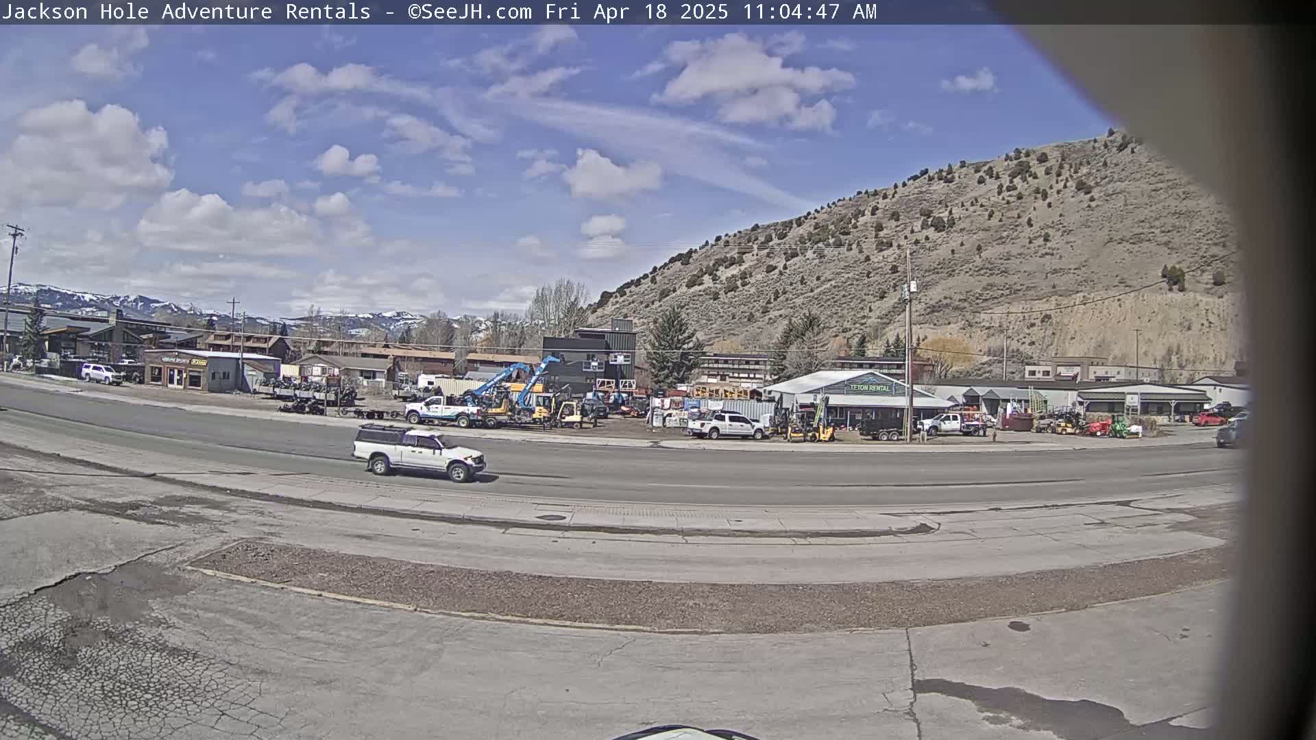 A partly cloudy day reveals a street scene with various vehicles, including a pickup truck, near a collection of buildings and equipment rentals against a backdrop of a mountainside.