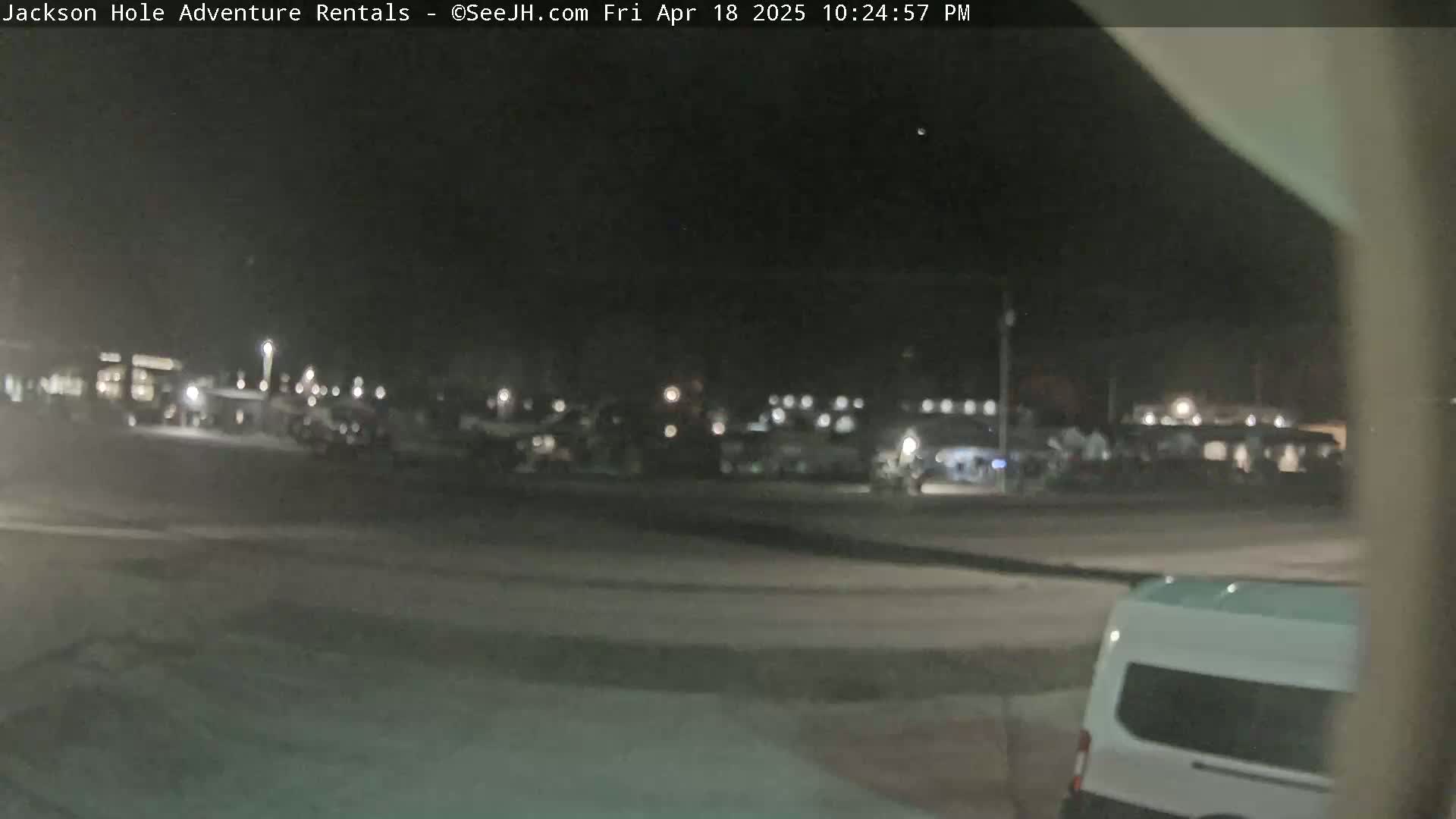 A nighttime, outdoor view shows a mostly empty parking lot with several vehicles parked and a white van in the foreground.