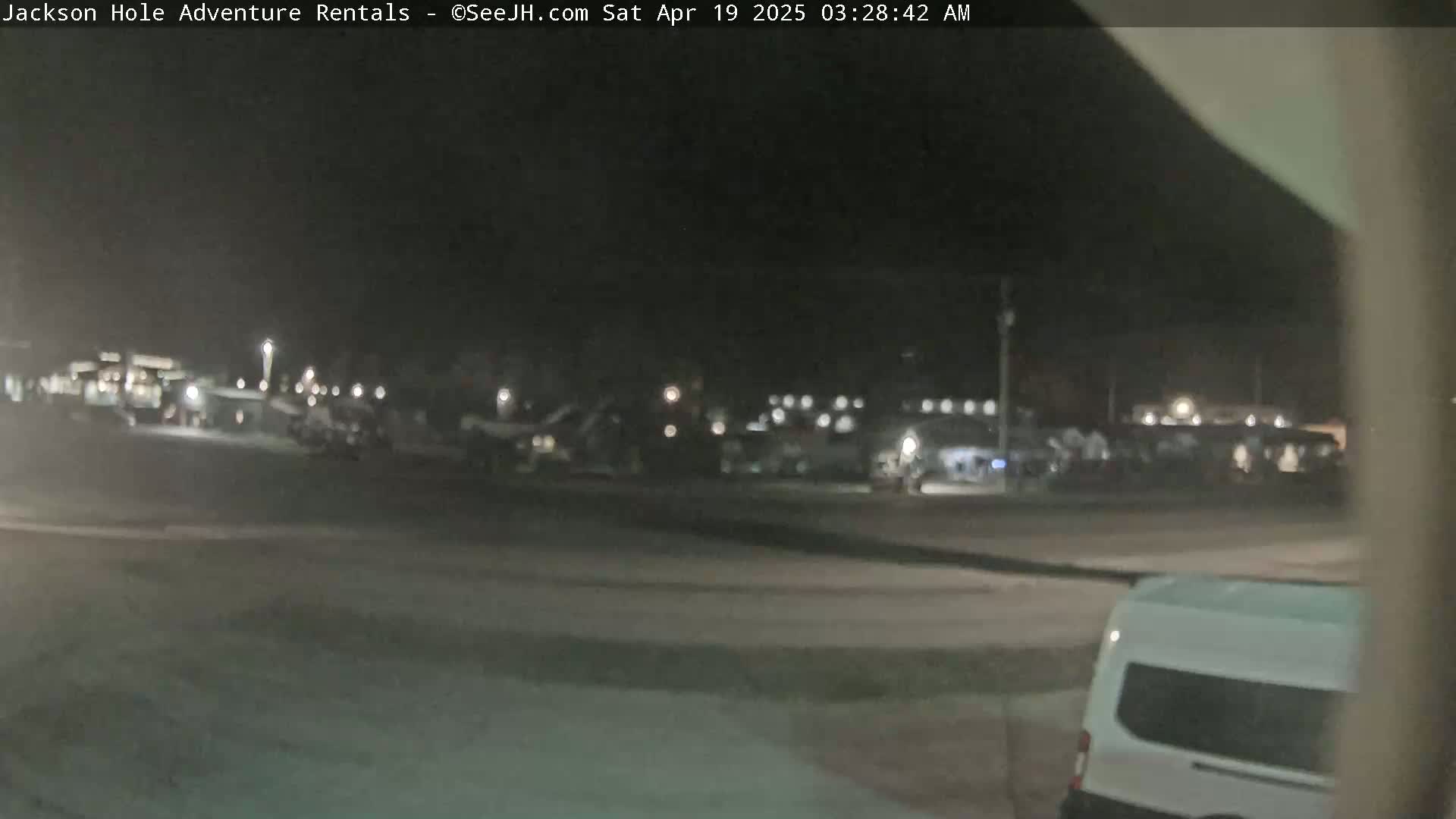 A nighttime view shows a mostly empty parking lot with several vehicles parked in the distance under a dark sky.