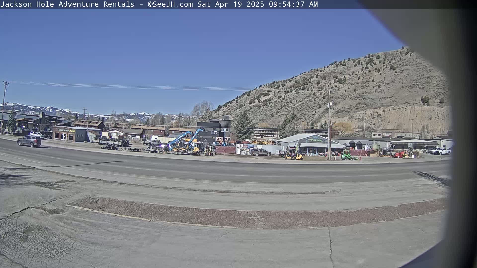 A sunny day reveals a street scene with several vehicles parked near rental businesses and equipment, backed by a hill and snow-capped mountains in the distance.