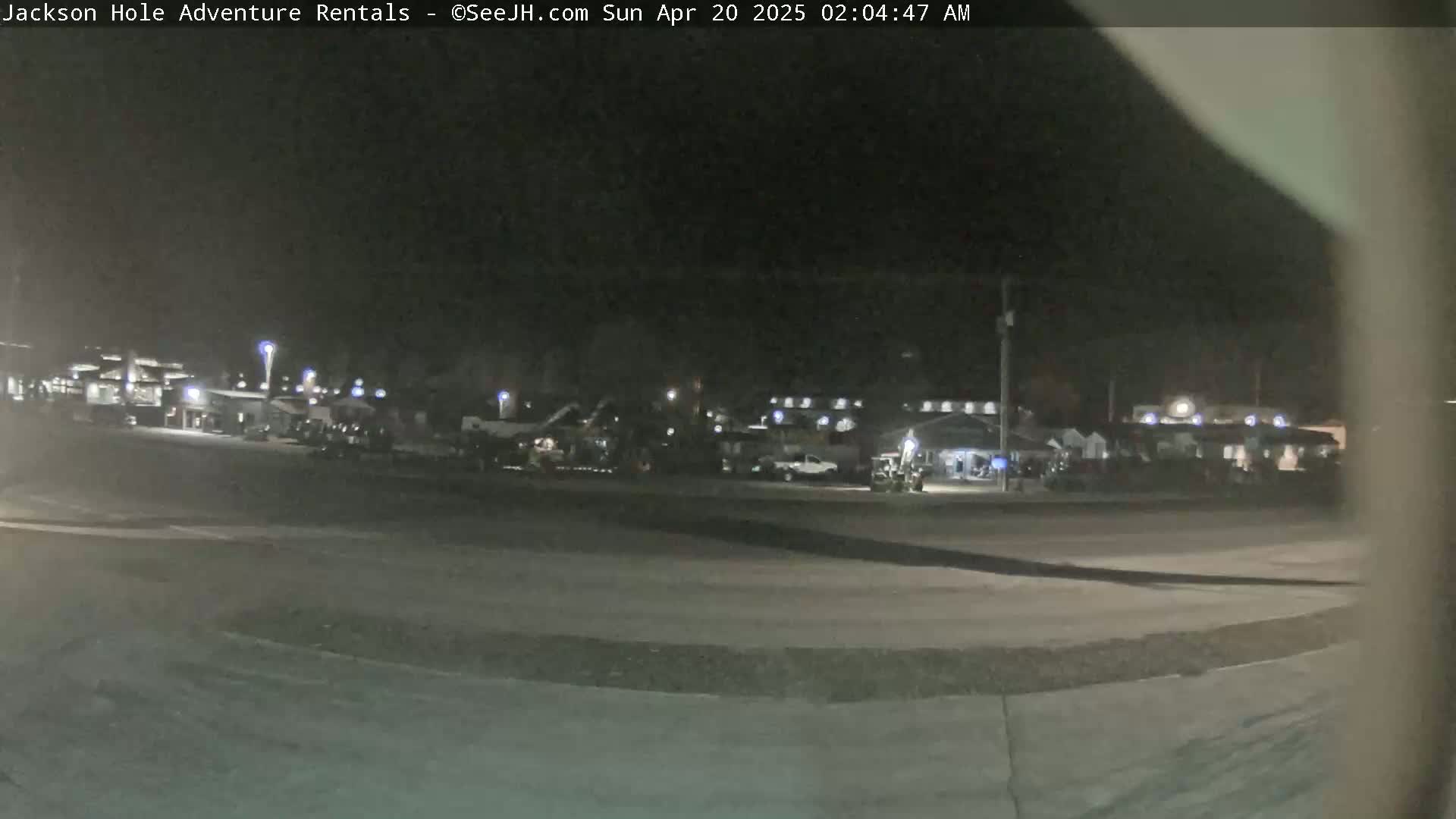 A nighttime, outdoor view of a parking lot with several buildings and vehicles under dim lighting.