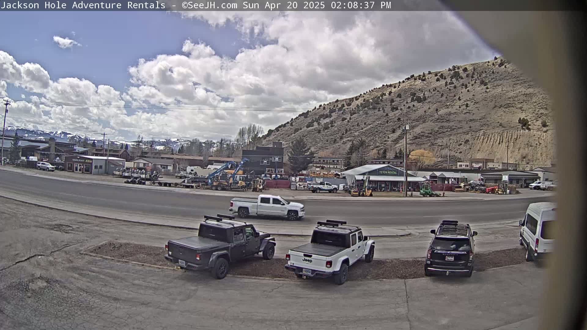 A partly cloudy day reveals a street scene with several parked pickup trucks and SUVs in front of a row of buildings, including a rental business, against a backdrop of a dry, hilly landscape.