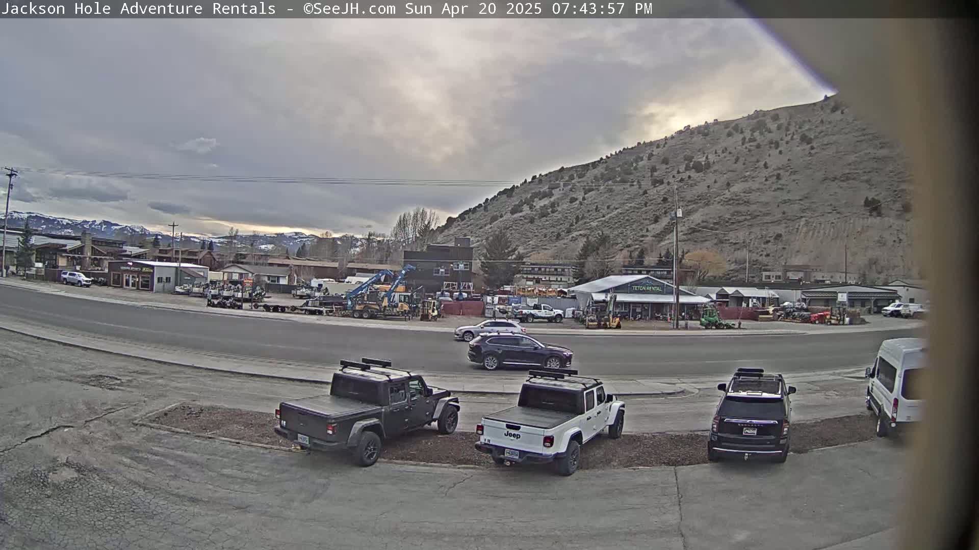 A mostly cloudy evening scene shows a parking lot with several SUVs and pickup trucks, in front of a row of buildings and a large hill in the background.