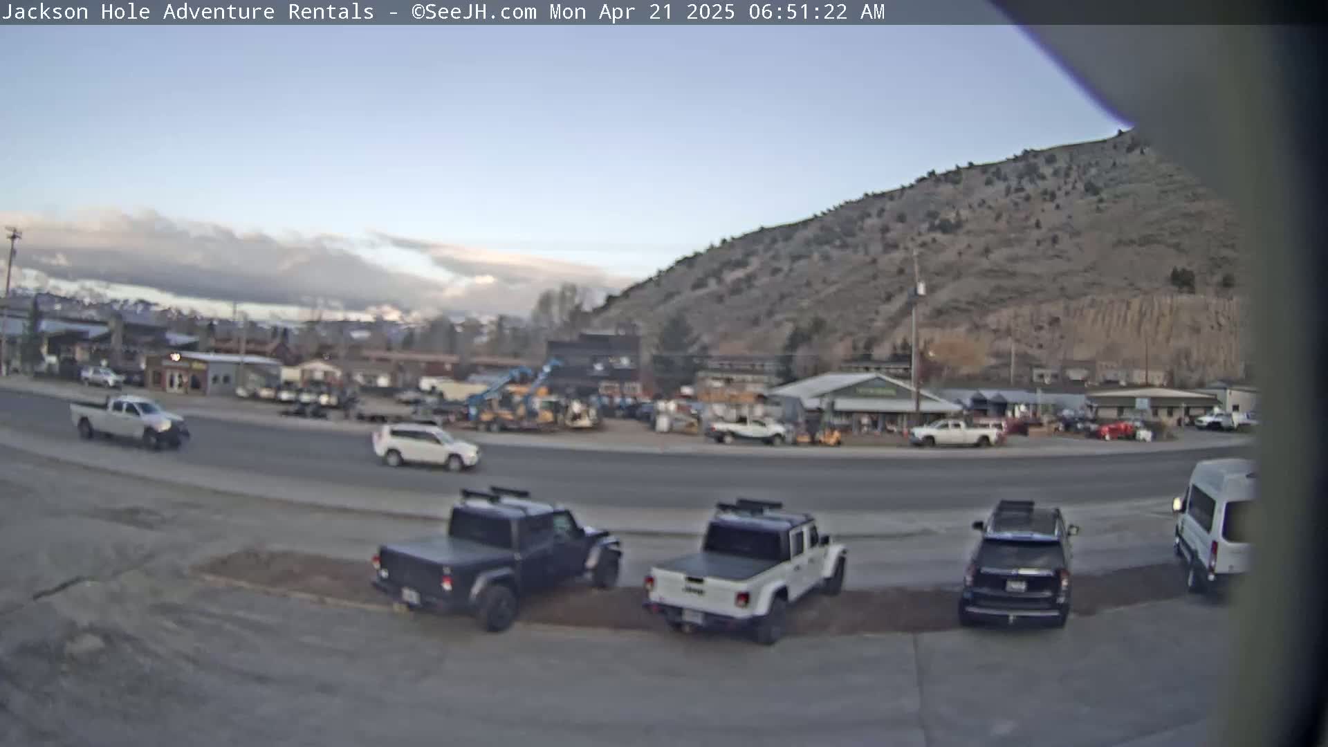 A mostly clear, early morning scene shows several vehicles parked and driving in a lot next to a road, with a hillside visible in the background.