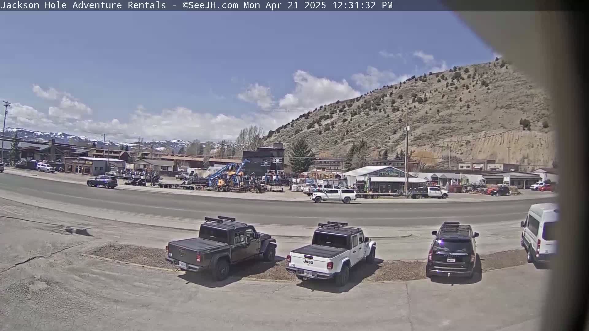 A sunny day reveals a street scene with several parked trucks and SUVs in front of a row of buildings, backed by a large, arid hill.
