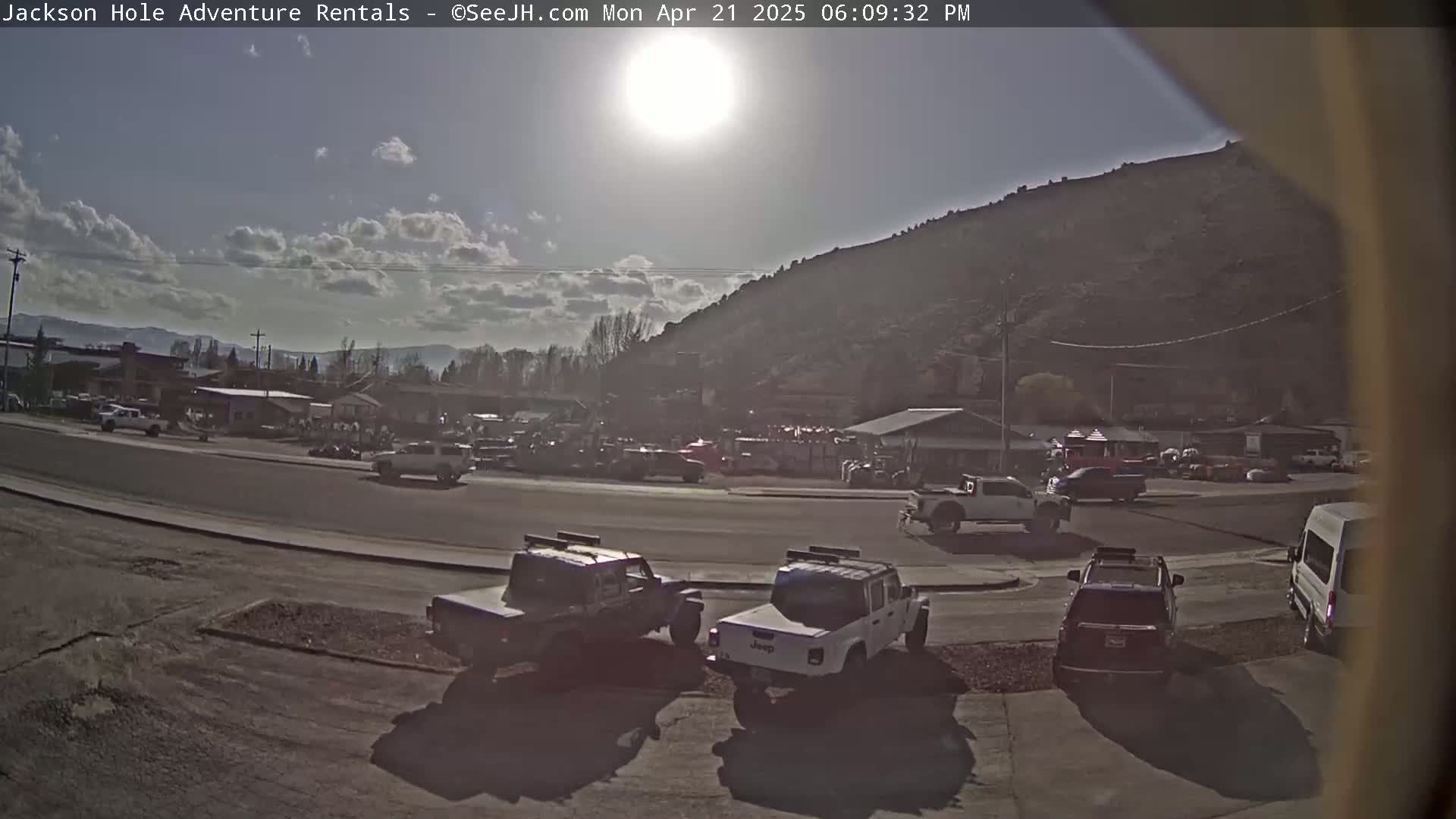A sunny day reveals a parking lot with several vehicles, a few buildings, and a large hill in the background.
