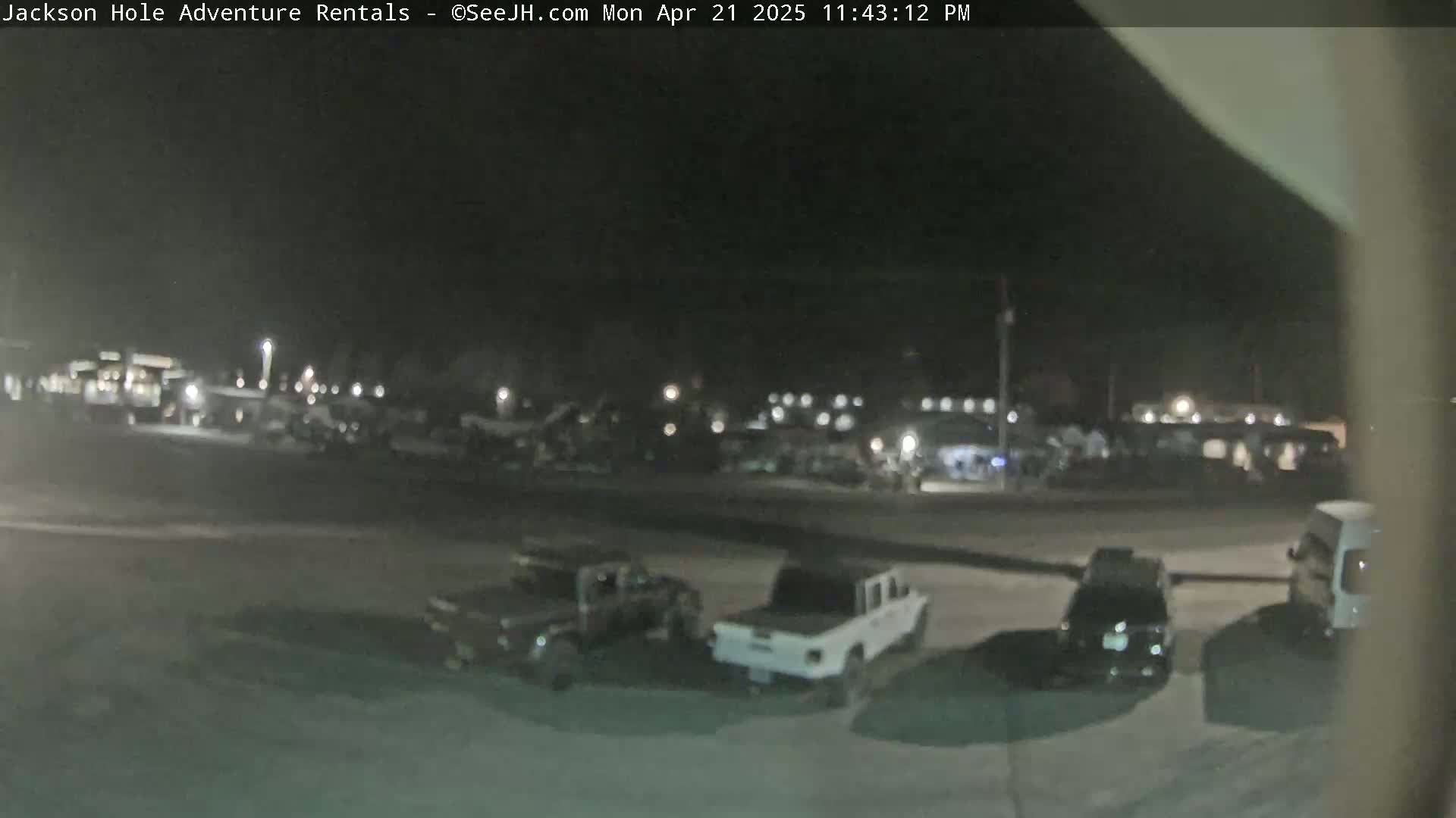 A nighttime, outdoor scene shows a parking lot with several vehicles, including trucks and SUVs, and some indistinct buildings in the background.