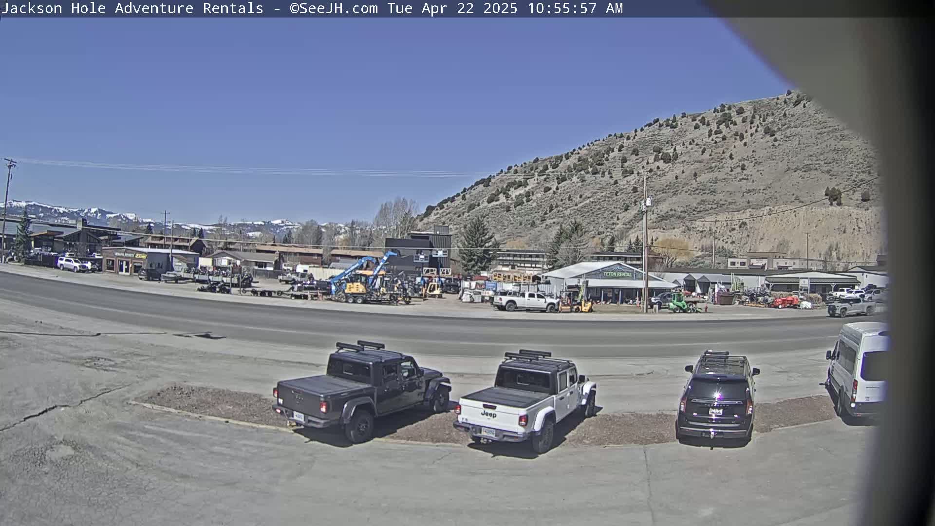 A sunny day reveals a parking lot with several vehicles, including two Jeeps, in front of a row of businesses and a large, barren hill in the background.