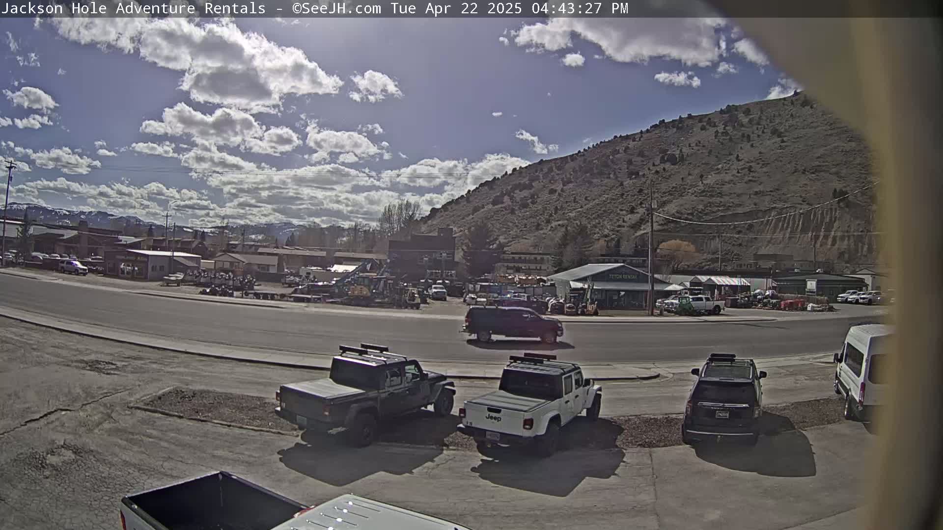 A sunny day reveals a parking lot with several Jeeps and other vehicles in front of a row of buildings and a large hill, under a partly cloudy sky.
