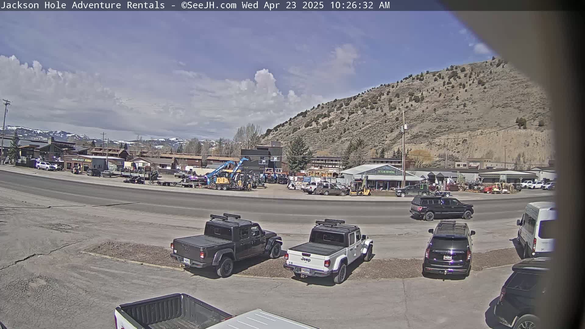 A partly cloudy day reveals a street scene with several parked trucks and SUVs near a business, backed by a mountainside.