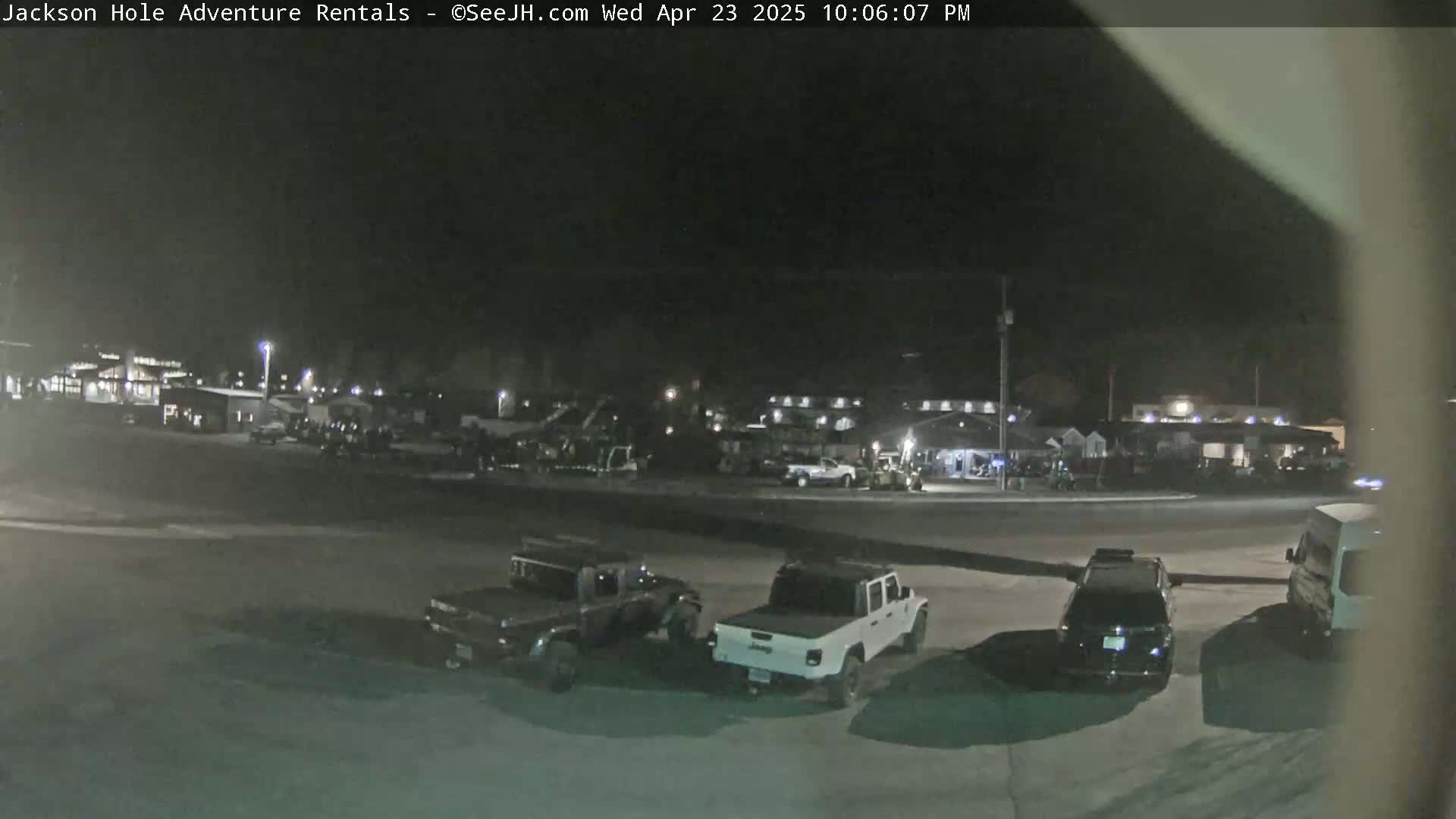 A nighttime scene shows several vehicles parked in a lot in front of a building complex under a dark sky.