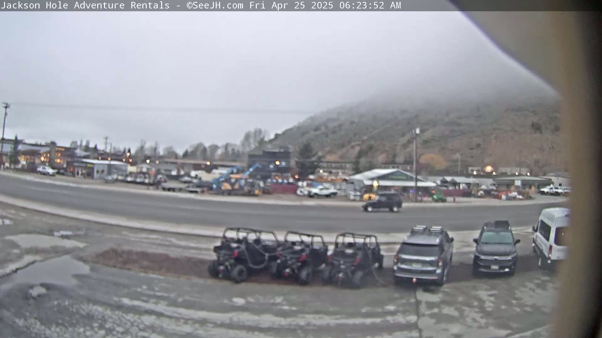 A foggy, overcast morning scene shows a street with several parked all-terrain vehicles, cars, and a van in front of a building and other vehicles and equipment further back.
