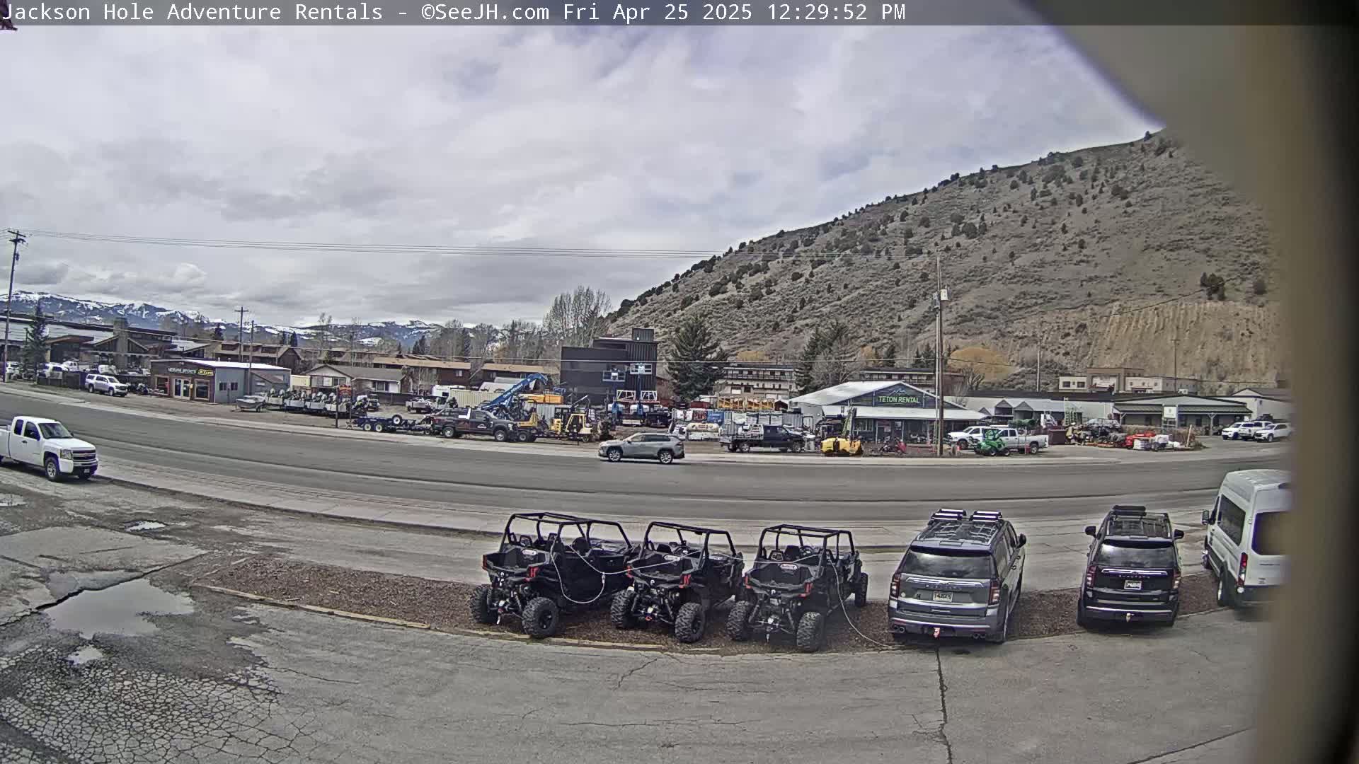 A street scene on an overcast day shows several parked off-road vehicles in front of a row of businesses and a hill in the background.