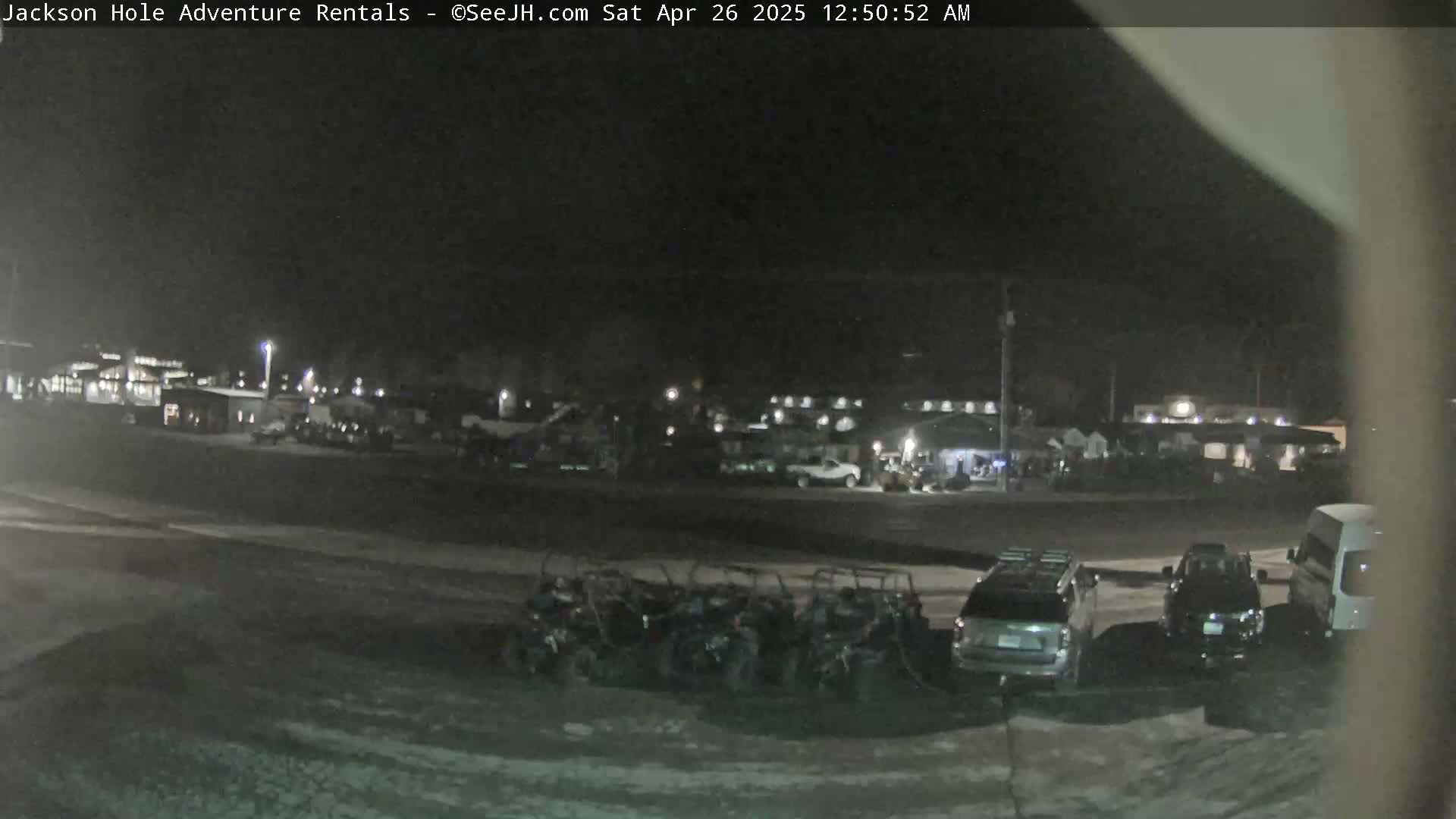 A nighttime scene shows a parking lot with several cars and all-terrain vehicles parked near a building complex under dark, clear skies.