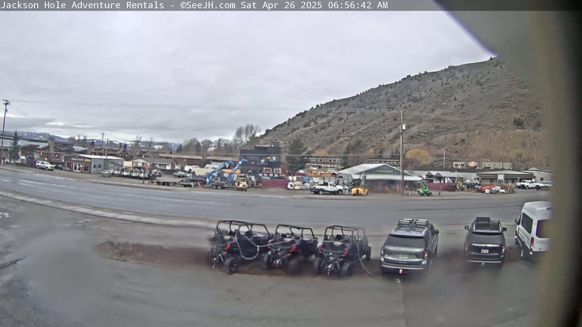 Several all-terrain vehicles are parked in a lot near a road, in front of various buildings and businesses, under an overcast sky.