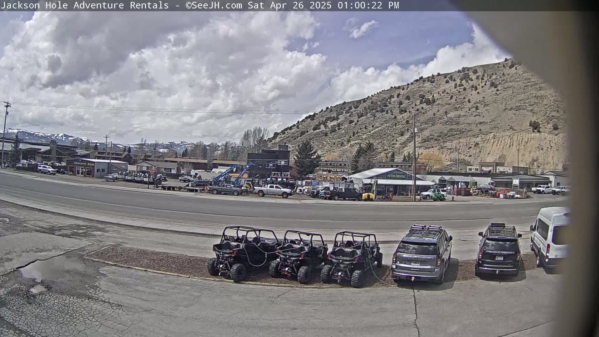 A partly cloudy day reveals a street scene with several parked SUVs and ATVs in front of a rental business, and a mountainside in the background.