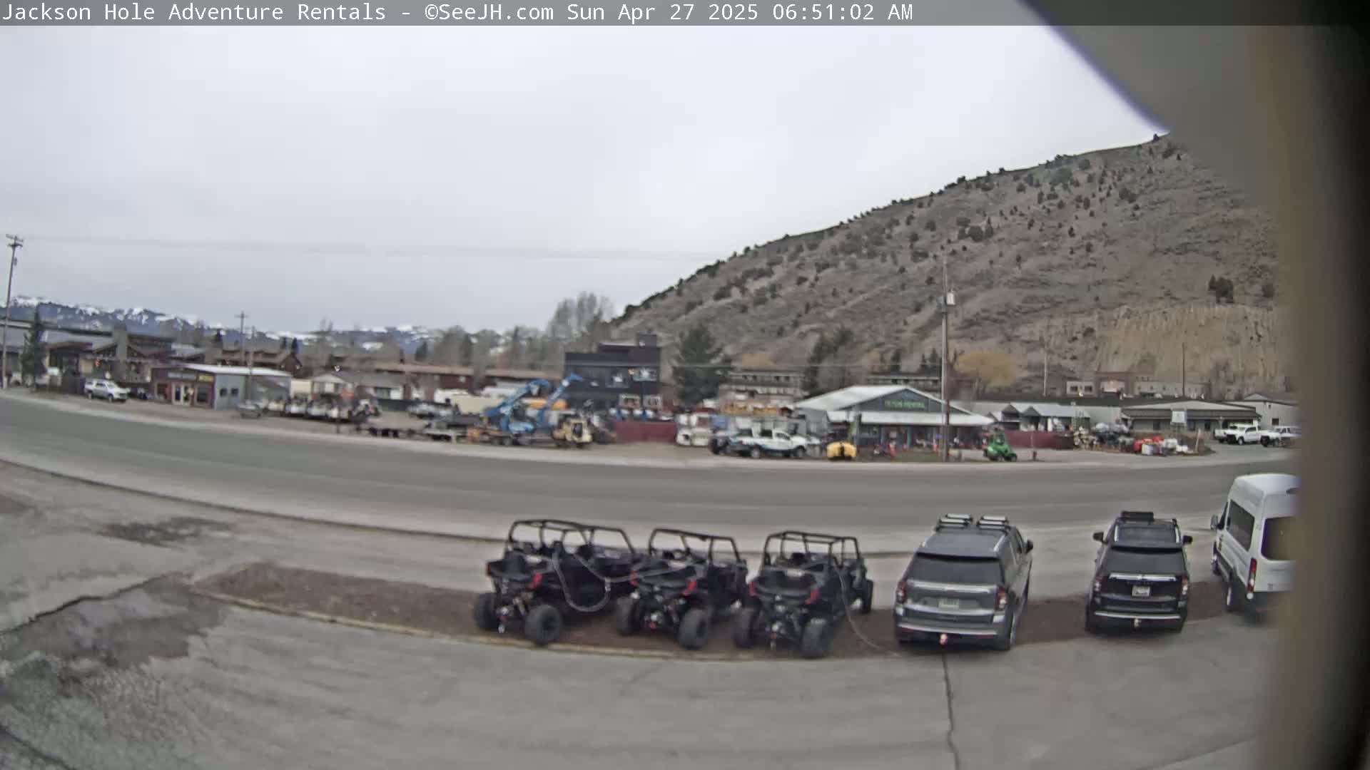 Several off-road vehicles are parked in a lot next to a street in front of buildings and a hill, under an overcast sky.