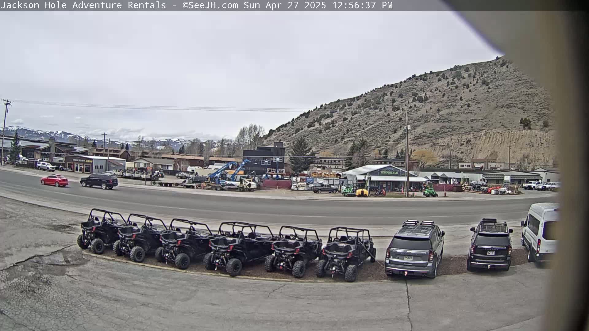 A row of black all-terrain vehicles is parked in a lot next to a road with cars and trucks, in front of a hill under an overcast sky.