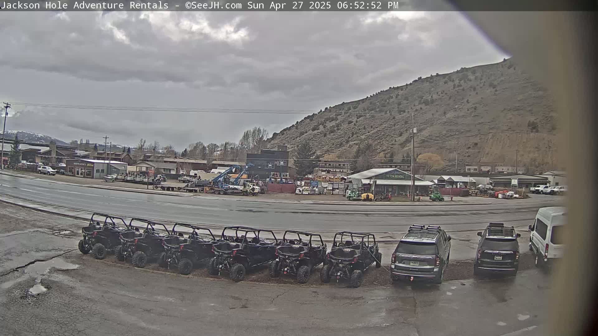 A row of all-terrain vehicles is parked in a lot next to a street, in front of several buildings and a hill, under a cloudy sky.